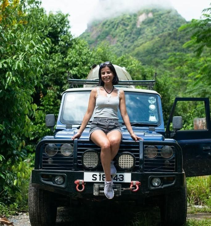 En kvinde sidder på kølerhjelmen af en åben Land Rover Defender med en grøn bjergside bagved