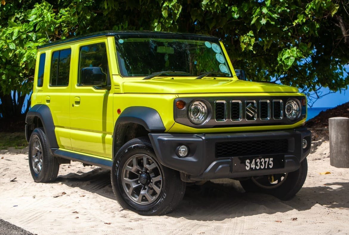 En gul Suzuki Jimny med fem døre holder i sandet under takamakatræer